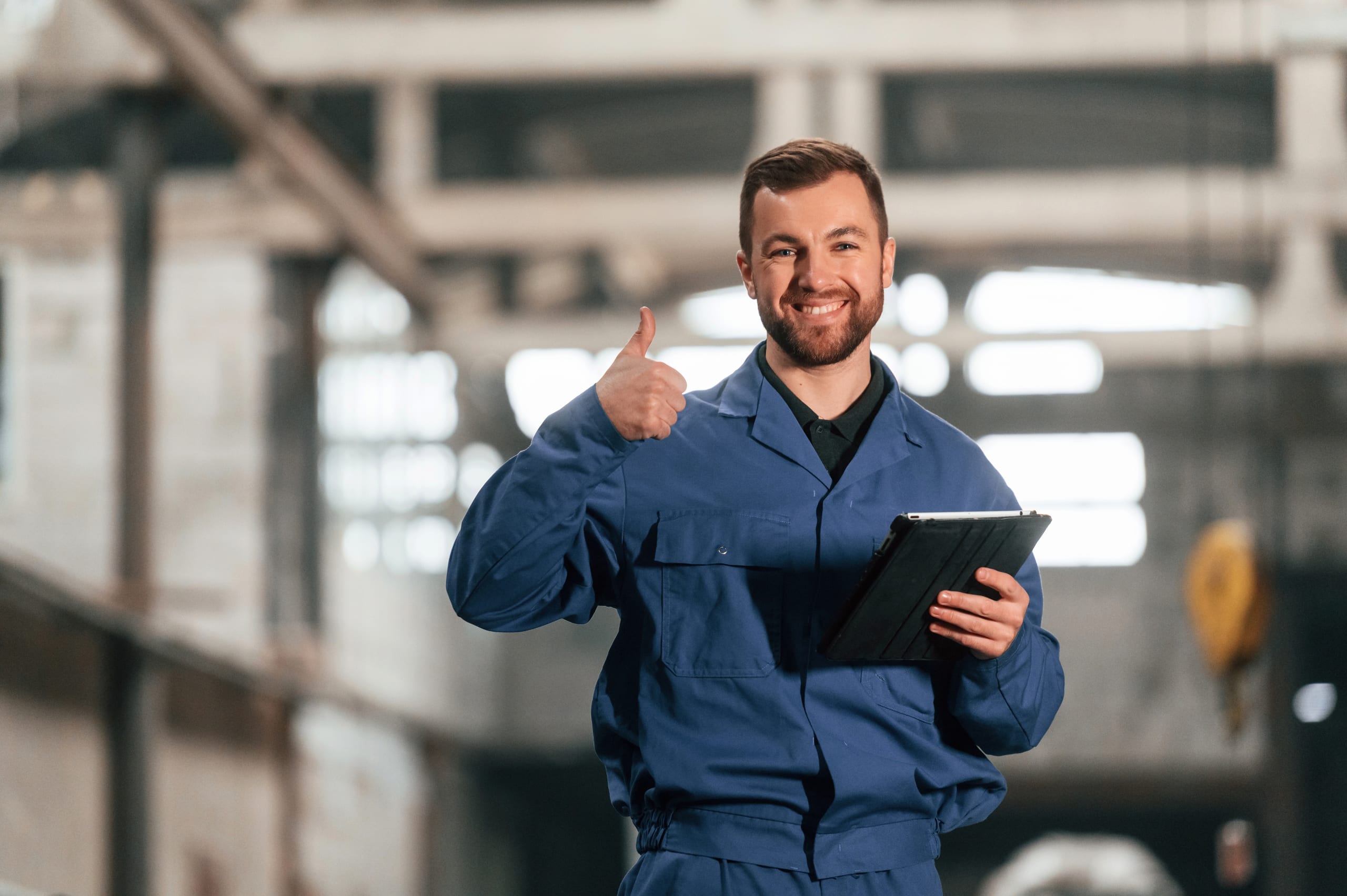 Smiling worker in blue overalls giving a thumbs up while holding a tablet